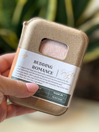 A hand holding Budding Romance Sea Salt and Clay Spa Soap Bar wrapped in a kraft paper sleeve with a clear window showing pink bath salts