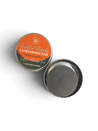 Open tin of RoseMint Rhapsody Conditioner Bar showing the solid black bar, next to the closed tin with its front label